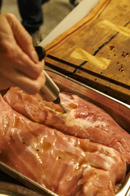 Eine Hand hält eine Marinadenspritze und injiziert eine Flüssigkeit in ein rohes Schweinefilet, das sich in einer silbernen Metallform befindet. Auf dem Filet sind bereits einige Tropfen der Marinade verteilt. Im Hintergrund befindet sich ein Holzbrett.